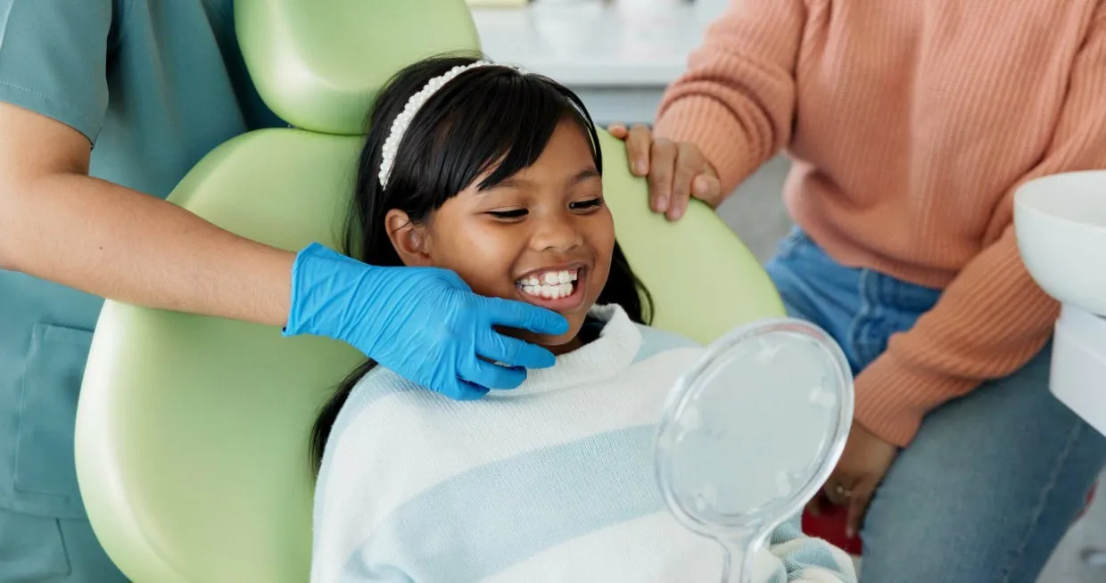 Young girl smiling in a dental chair, receiving care from a dentist wearing blue gloves, with a parent nearby, promoting pediatric dental services at Kids Care Dental and Orthodontics in Hallandale Beach, FL.